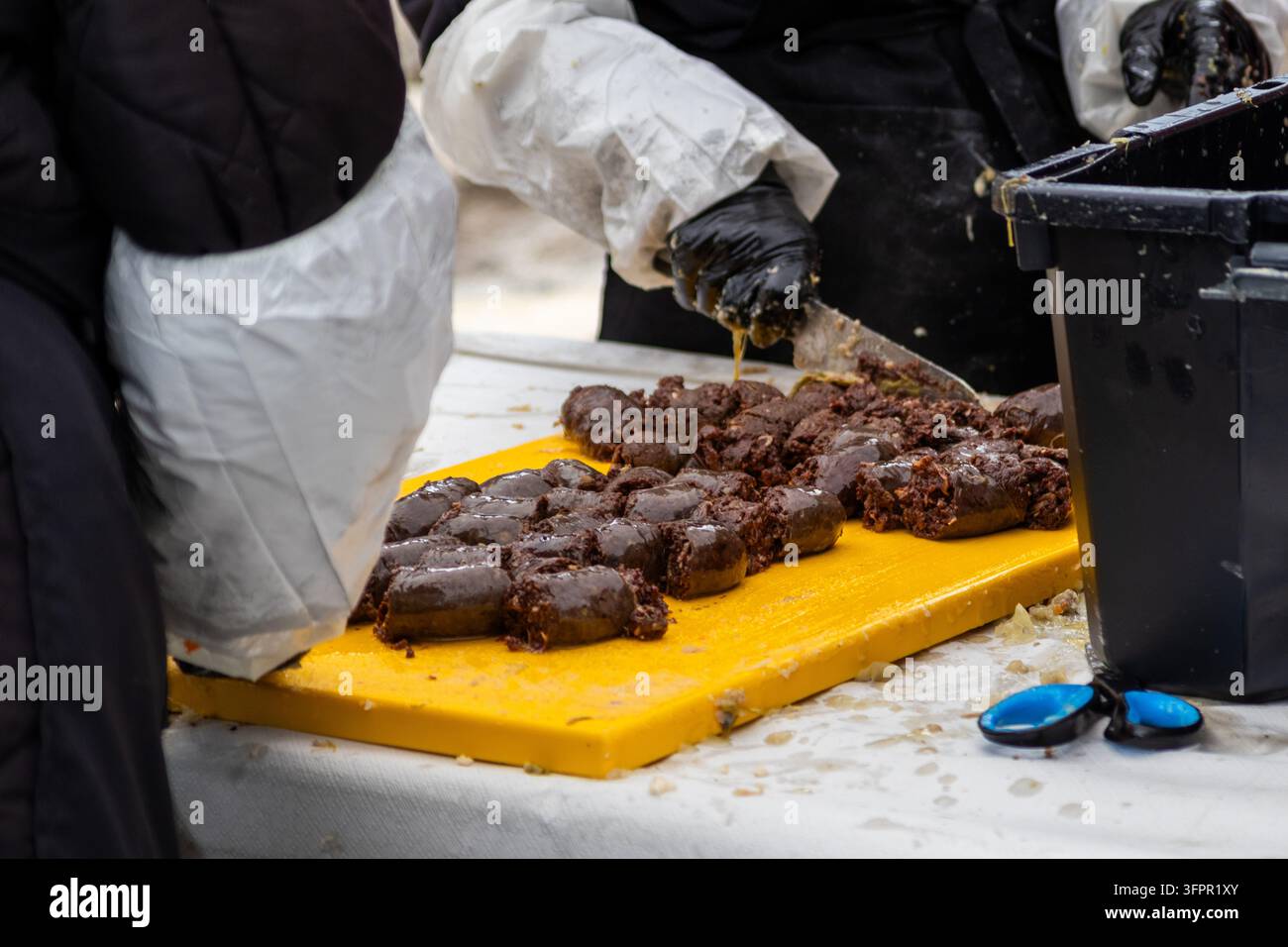 Mani indossando guanti neri che tagliano la tradizionale salsiccia nera butifarra su un tagliere giallo durante la preparazione dello stufato escudella Foto Stock