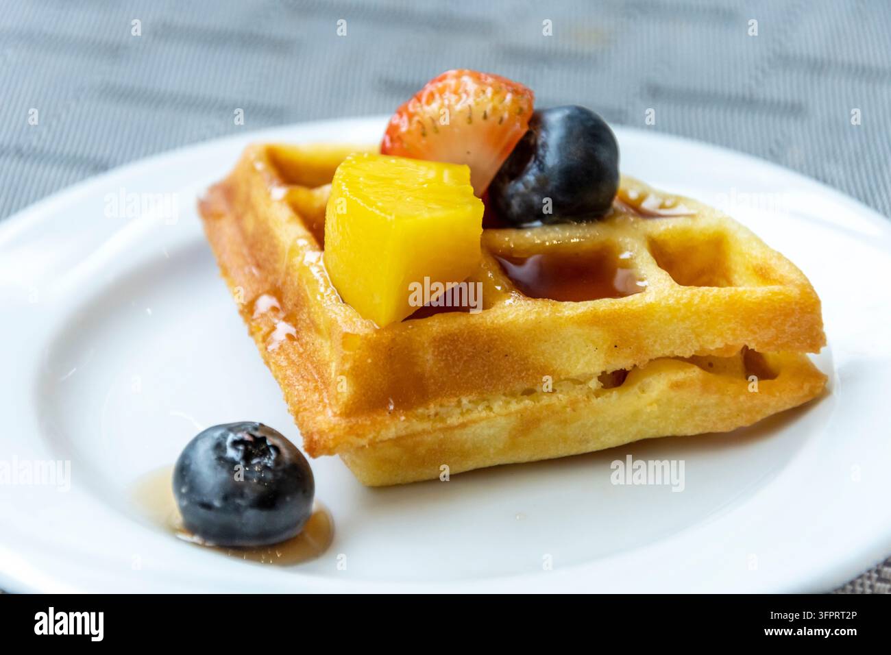 Una singola waffle quadrata spruzzata con sciroppo dolce e guarnita con pezzi di frutta tropicale vivaci per una deliziosa presentazione di colazione o dessert Foto Stock