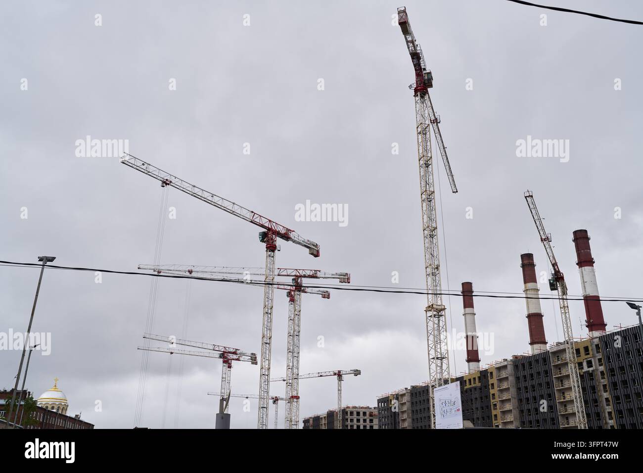 Le gru per torre si ergono sopra edifici scheletrici in un cantiere edile urbano affollato, con camini e cielo grigio e coperto sullo sfondo Foto Stock