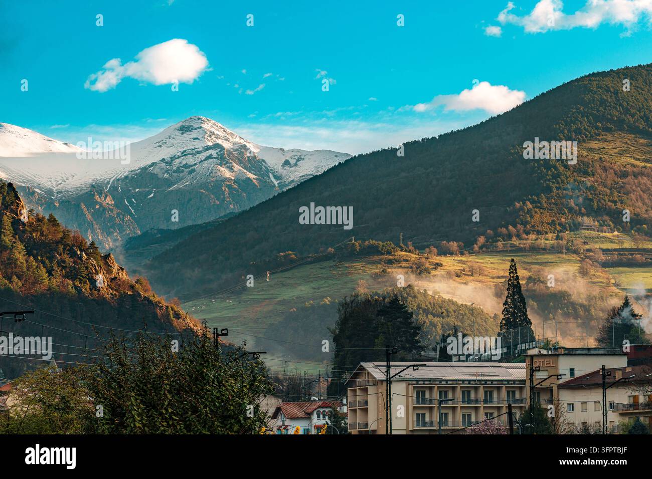 Un picco pirenaico coperto di neve si erge sopra una valle boscosa, prati verdi e nebbia che sale, con un hotel di villaggio e un alto abete a Ribes de Fresser Foto Stock