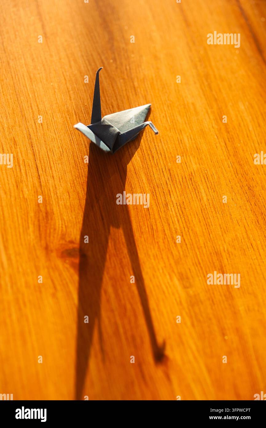 Piccola gru di origami grigia su un tavolo di legno arancione illuminato dal sole, che proietta un'ombra insolitamente lunga e nitida sulla venatura calda del legno Foto Stock