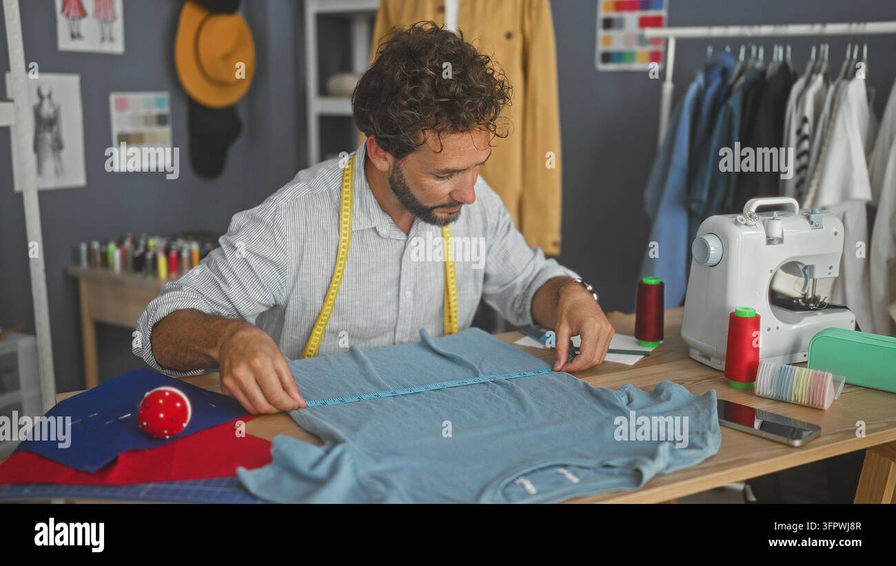 Sarto uomo che misura una camicia blu con il metro, mano che si allunga per lisciare il tessuto e posizionare le puntine accanto alla macchina da cucire sul tavolo da lavoro in studio; artigianalità Foto Stock