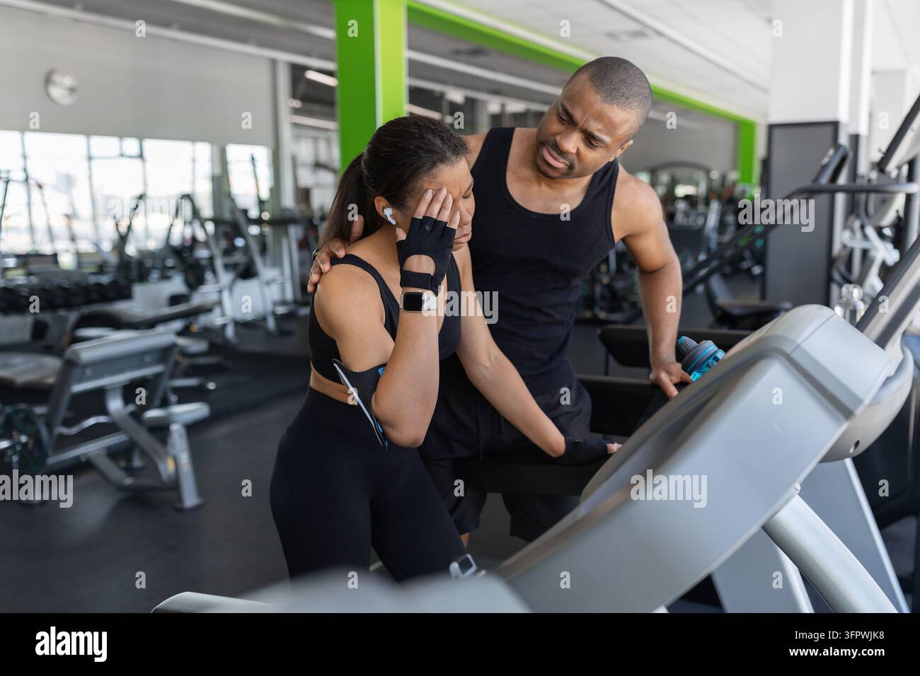 Uomo nero preoccupato che consola una donna che avverte disagio durante l'esercizio sul tapis roulant in palestra Foto Stock