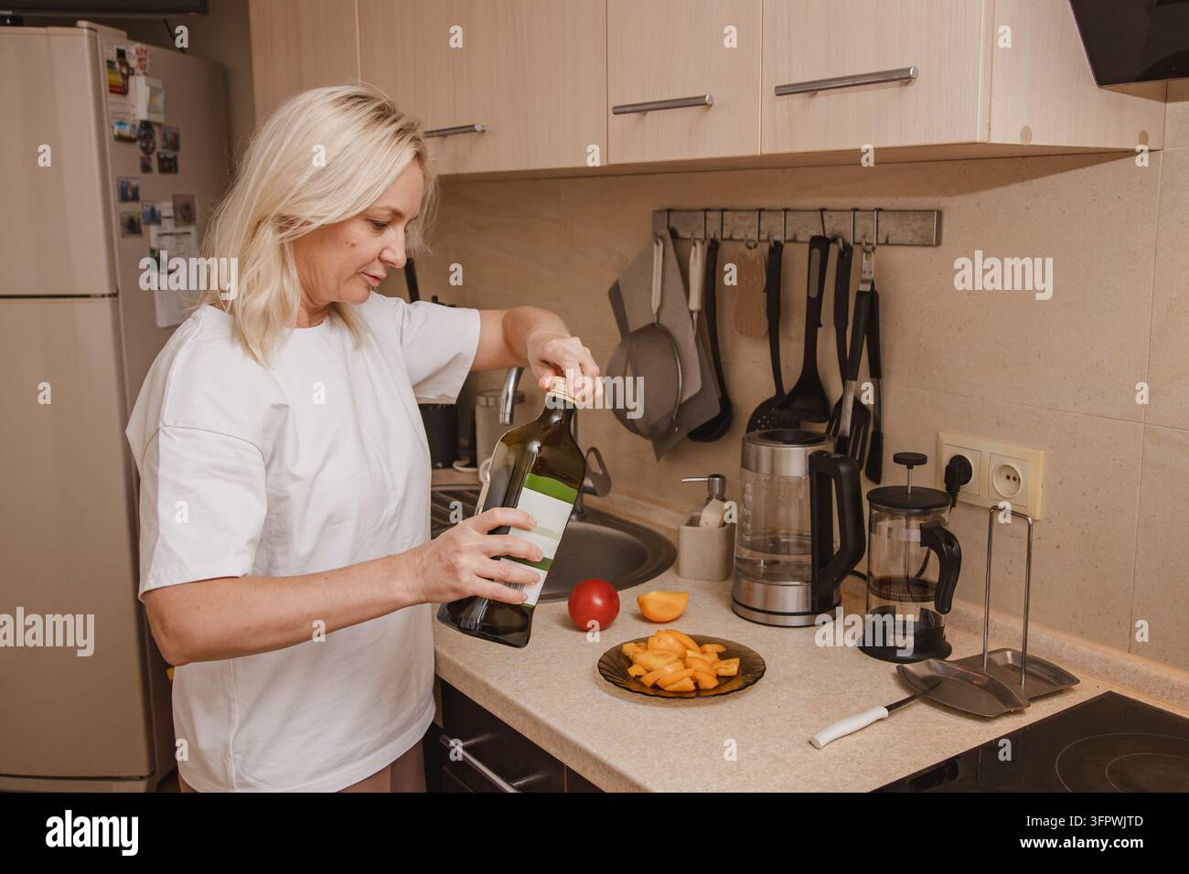 Una donna di mezza età è in piedi nella sua cucina e versa olio su rossi affettati Foto Stock