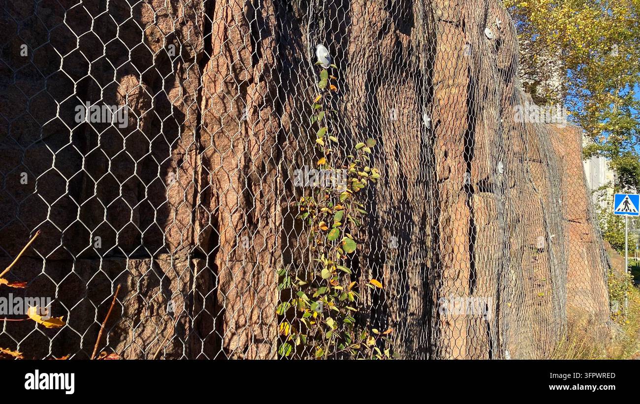 Muro di sostegno in pietra coperto da una rete protettiva per impedire la caduta di pietre lungo il bordo stradale urbano Foto Stock