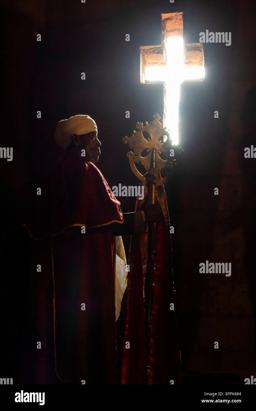 Un sacerdote in paramenti rossi e turbante bianco tiene una croce processionale dorata ornata mentre una luce intensa filtra attraverso un'apertura a forma di croce Foto Stock