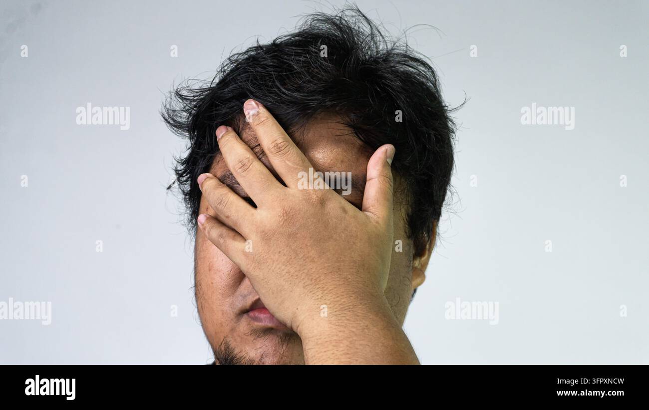 Un uomo asiatico adulto copre metà del suo viso con una mano in un gesto classico di frustrazione intensa, rimpianto o profonda stanchezza. L'individuo stan Foto Stock