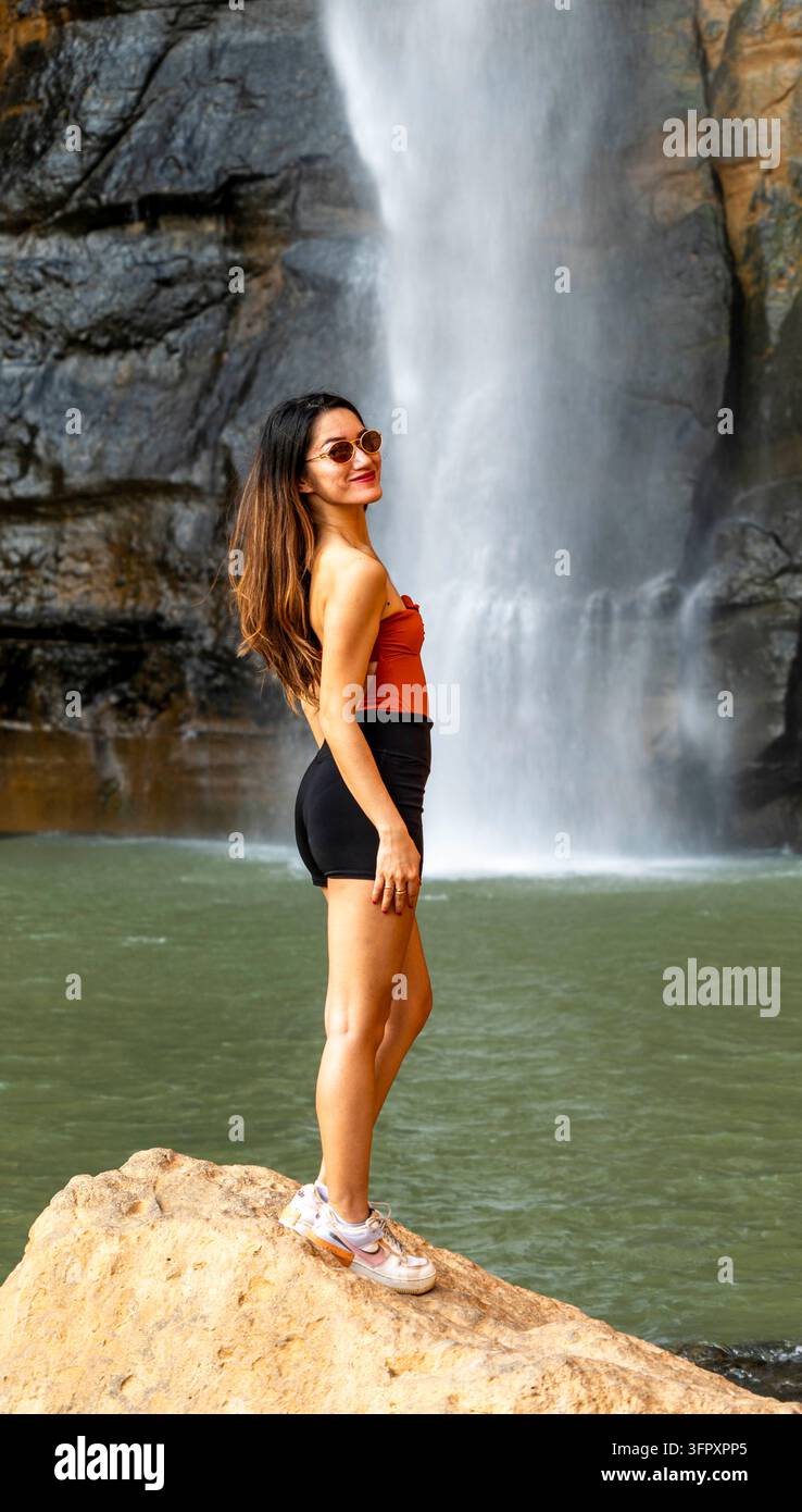 Giovane donna con occhiali da sole e abiti estivi casual in piedi su una roccia accanto a una potente cascata e a una piscina verde in un paesaggio naturale roccioso. Foto Stock