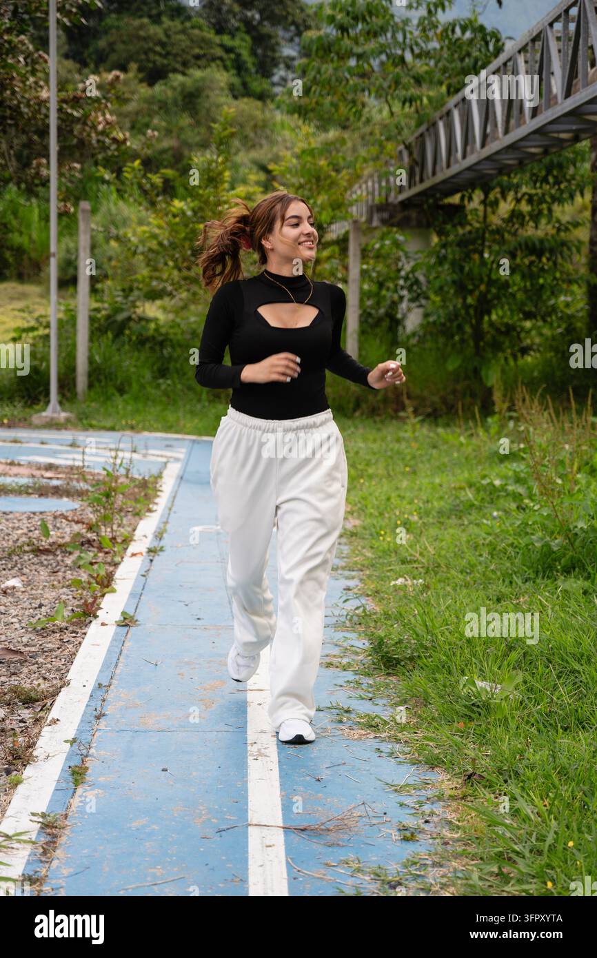 Giovane donna che corre su una pista esterna blu in un parco verde, fa jogging in abbigliamento sportivo per il fitness e il benessere, sorridente e concentrata sull'allenamento e la salute Foto Stock