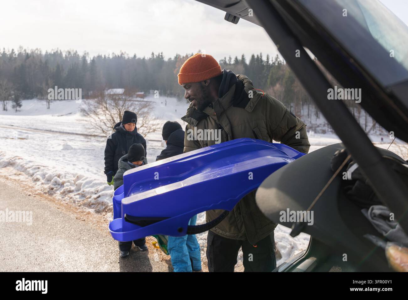 Padre afroamericano e i suoi tre figli prendono le loro slitte dall'auto, pronti per il divertimento in famiglia all'aperto e avventure invernali nella neve. Foto Stock