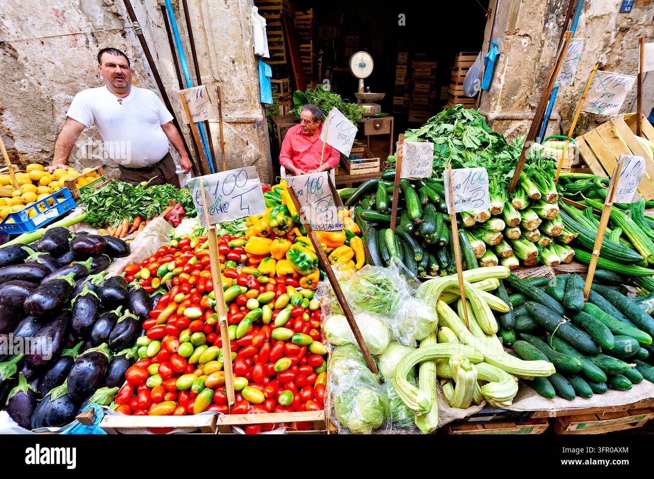 Due venditori con melanzane, pomodori, peperoni, zucchine e cartelli con prezzi in euro scritti a mano in un mercato alimentare all'aperto in Sicilia, Italia. Foto Stock
