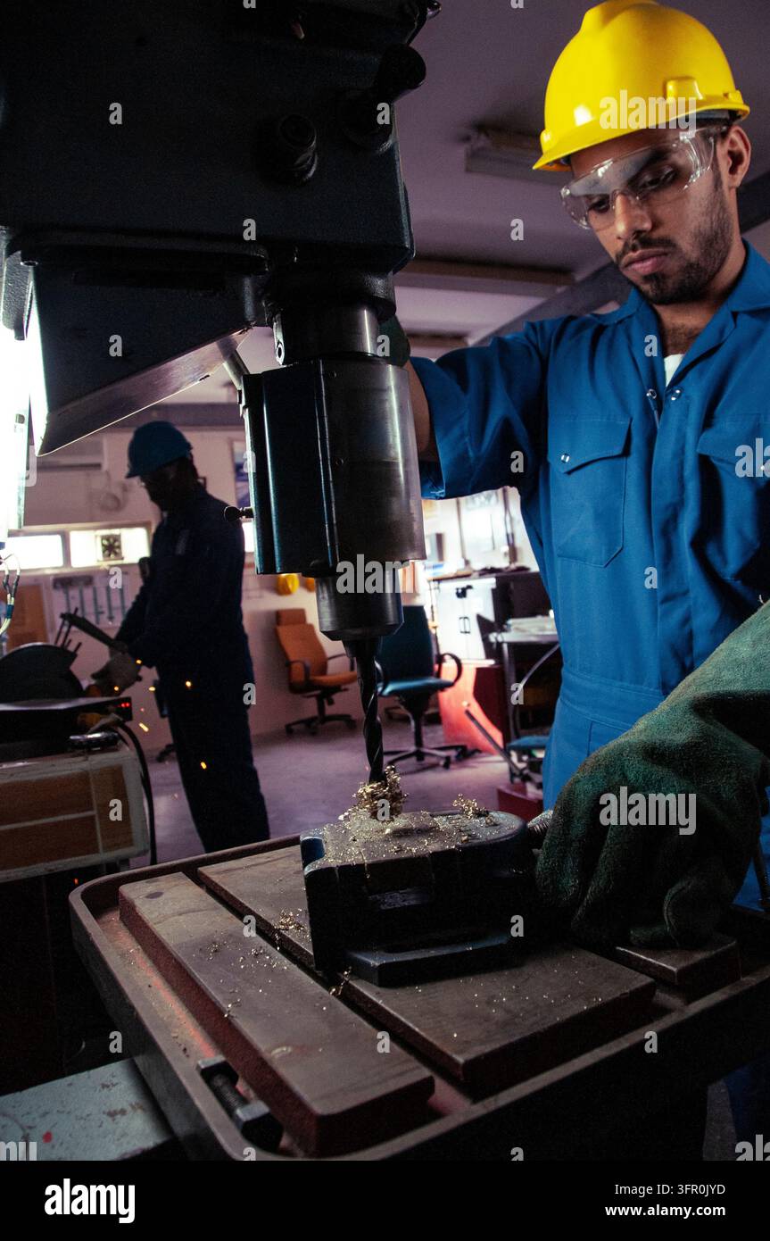 Lavoratore esperto in fabbrica indossa dispositivi protettivi e aziona una macchina di perforazione verticale mentre un collega lavora nell'area di sfondo Foto Stock