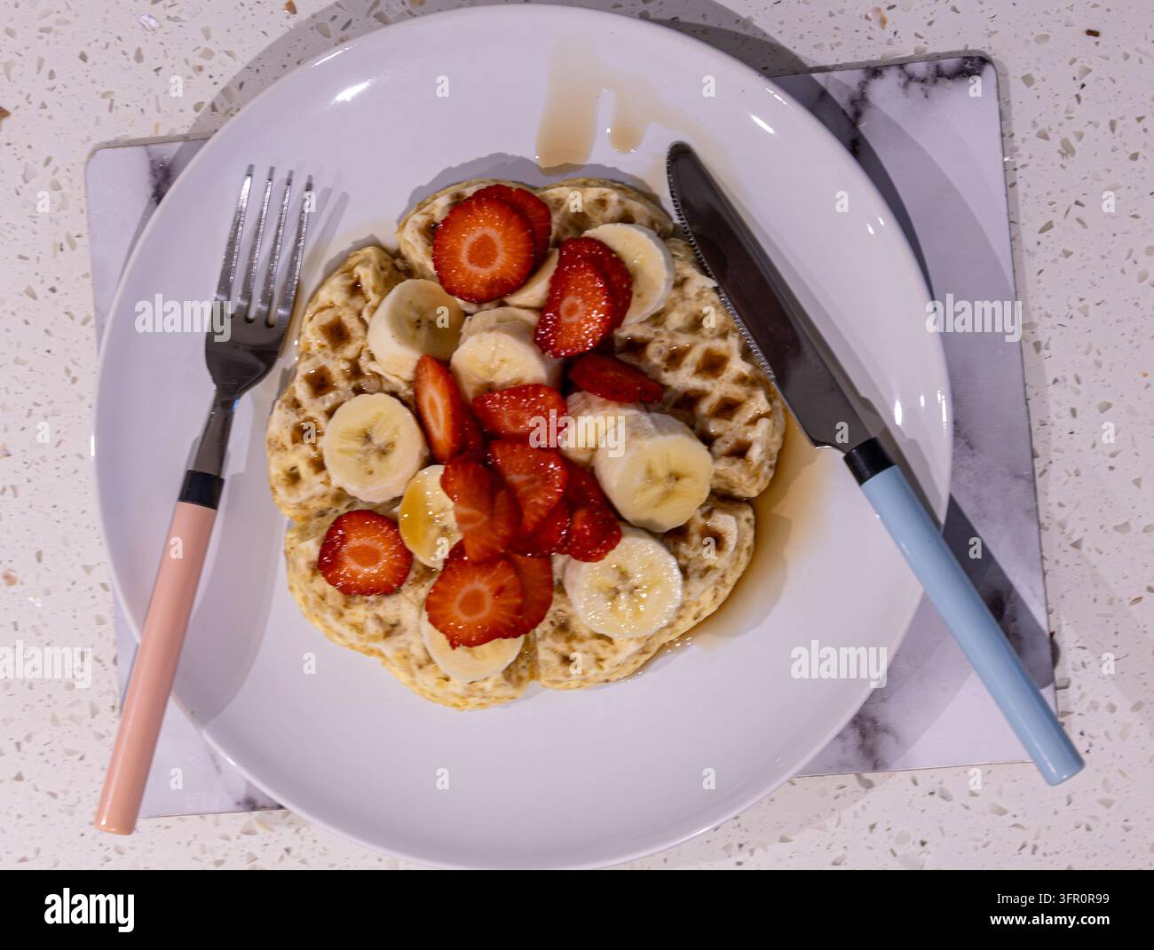 Vista dall'alto di waffle guarniti con fragole a fette, banana e sciroppo su un piatto bianco con manici di posate pastello su un piano di lavoro macchiato Foto Stock