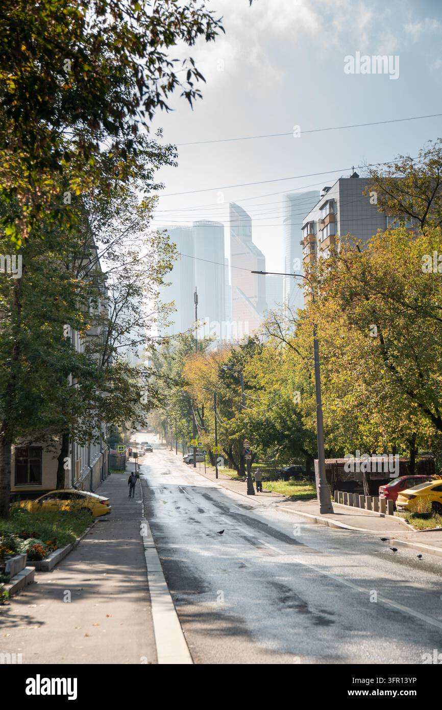 Mosca, Russia - 26 settembre 2026: i grattacieli di Moscow City si ergono in lontananza dietro una tranquilla strada residenziale con alberi autunnali. Sviluppo urbano Foto Stock