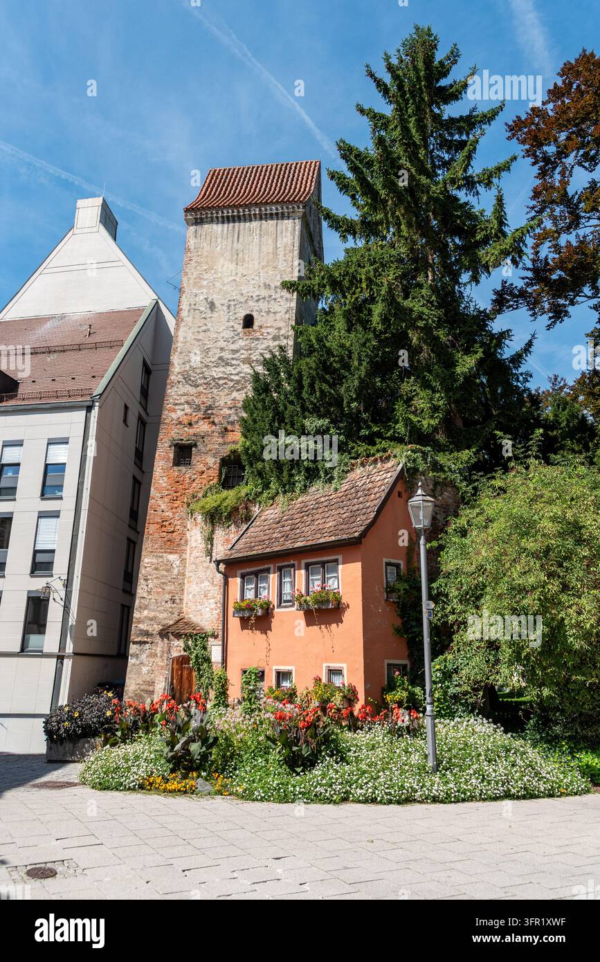 Scena idilliaca che mostra l'antica torre medievale Hexenturm dietro una piccola casa arancione decorata con vivaci fioriere alle finestre e un grande abete Foto Stock