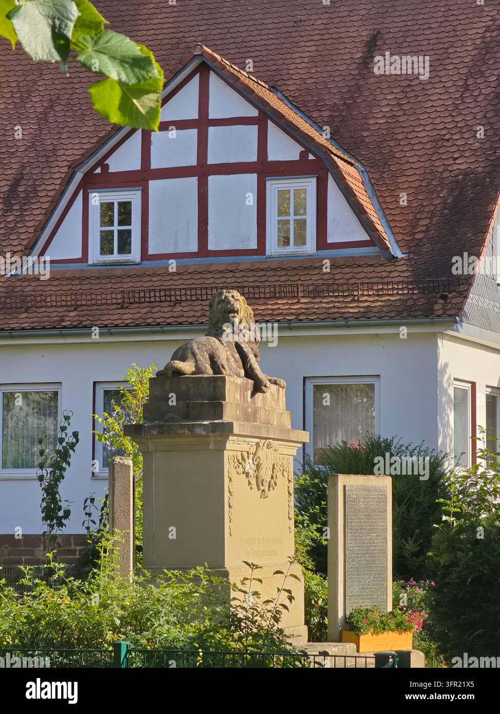 Monumento commemorativo in pietra a forma di leone in piedi all'aperto con un edificio tradizionale in legno a graticcio tedesco sullo sfondo in una giornata soleggiata. - Immagine stock catturata con smartphone