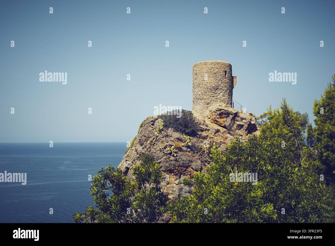 Una torre di guardia storica in pietra si erge in modo prominente su una scogliera aspra e rocciosa che si affaccia sul calmo mare Mediterraneo blu sotto un cielo limpido e luminoso a Mall Foto Stock