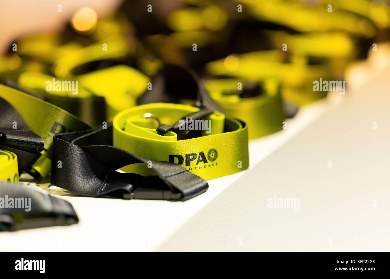 Amsterdam, Paesi Bassi – 11 settembre 2026: Lanyard verdi DPA Microfoni con clip neri si sovrappongono su una superficie bianca, con apparecchiature sfocate e wa Foto Stock