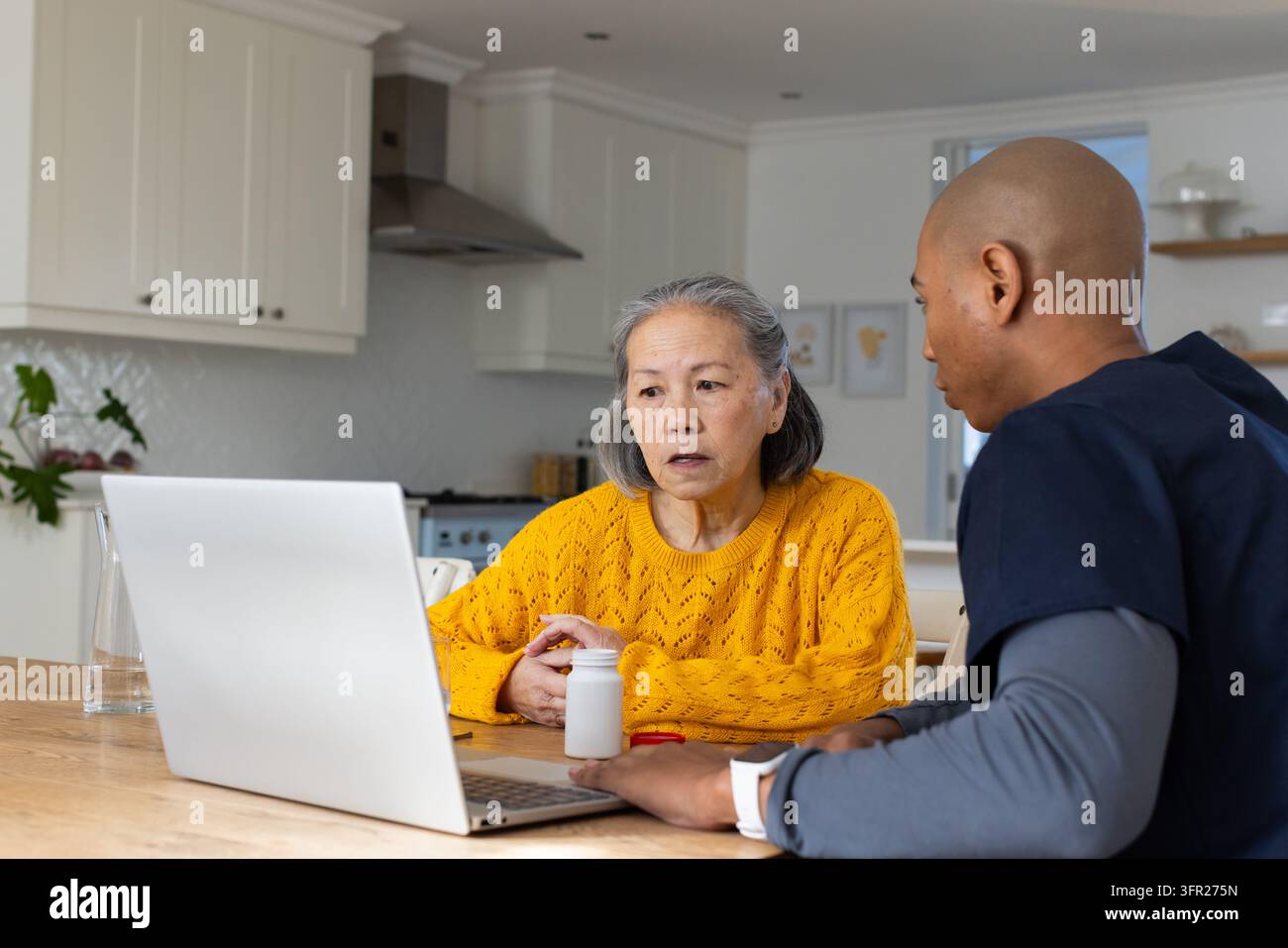 Diverse assistenti sanitari che aiutano un anziano asiatico in maglia gialla in cucina utilizzando un laptop e una bottiglia di pillole Foto Stock