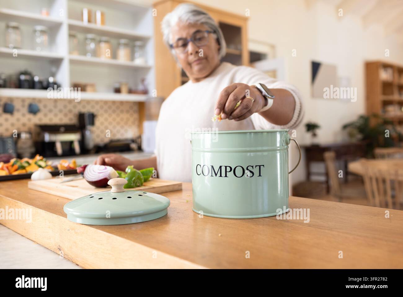 Donna anziana in maglione che lascia cadere avanzi di cibo in una scatola di menta etichettata COMPOST in cucina Foto Stock