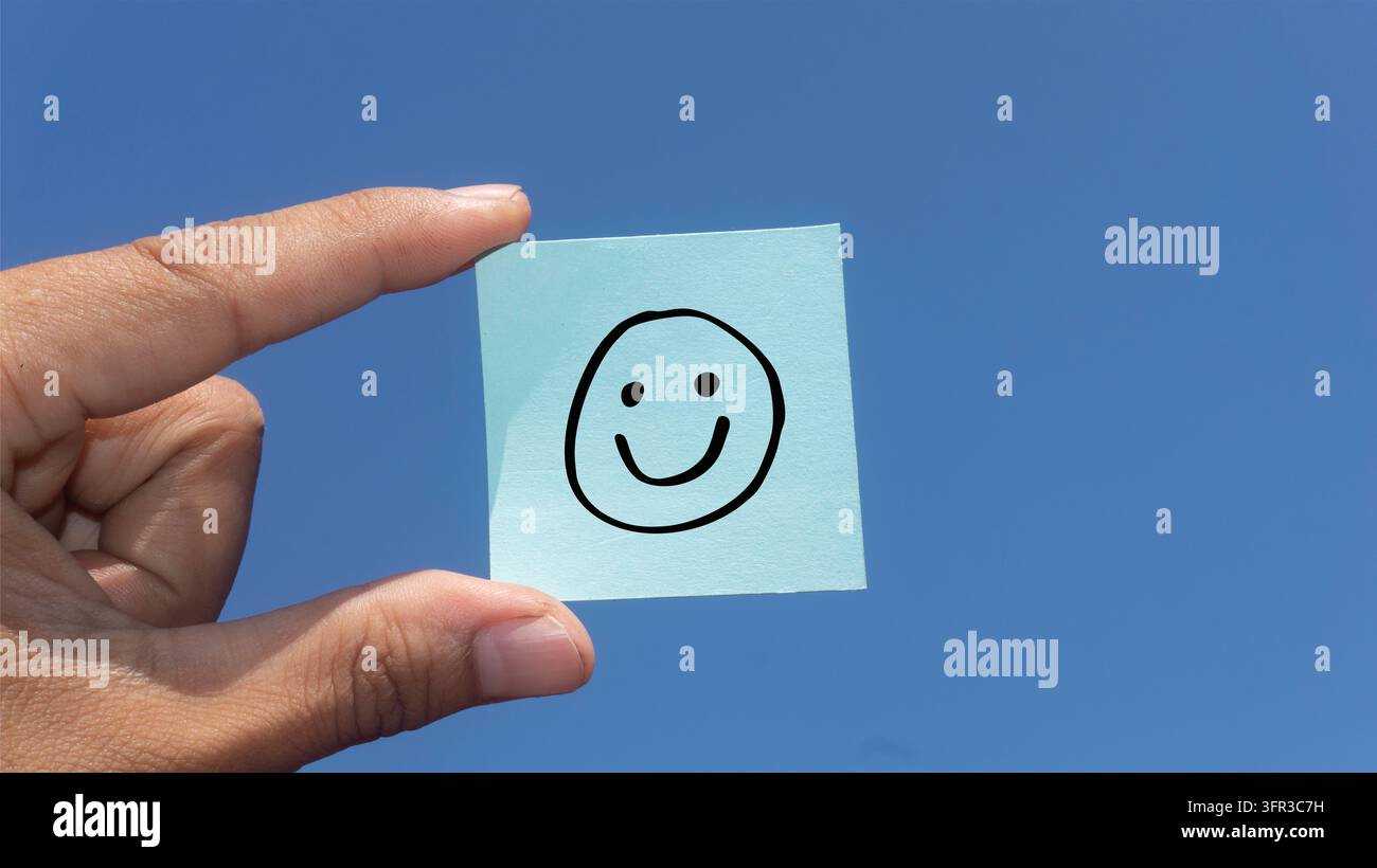 Mano che tiene un post-it con un viso sorridente su uno sfondo di cielo blu, rappresentante felicità, positività, soddisfazione, feedback del cliente Foto Stock