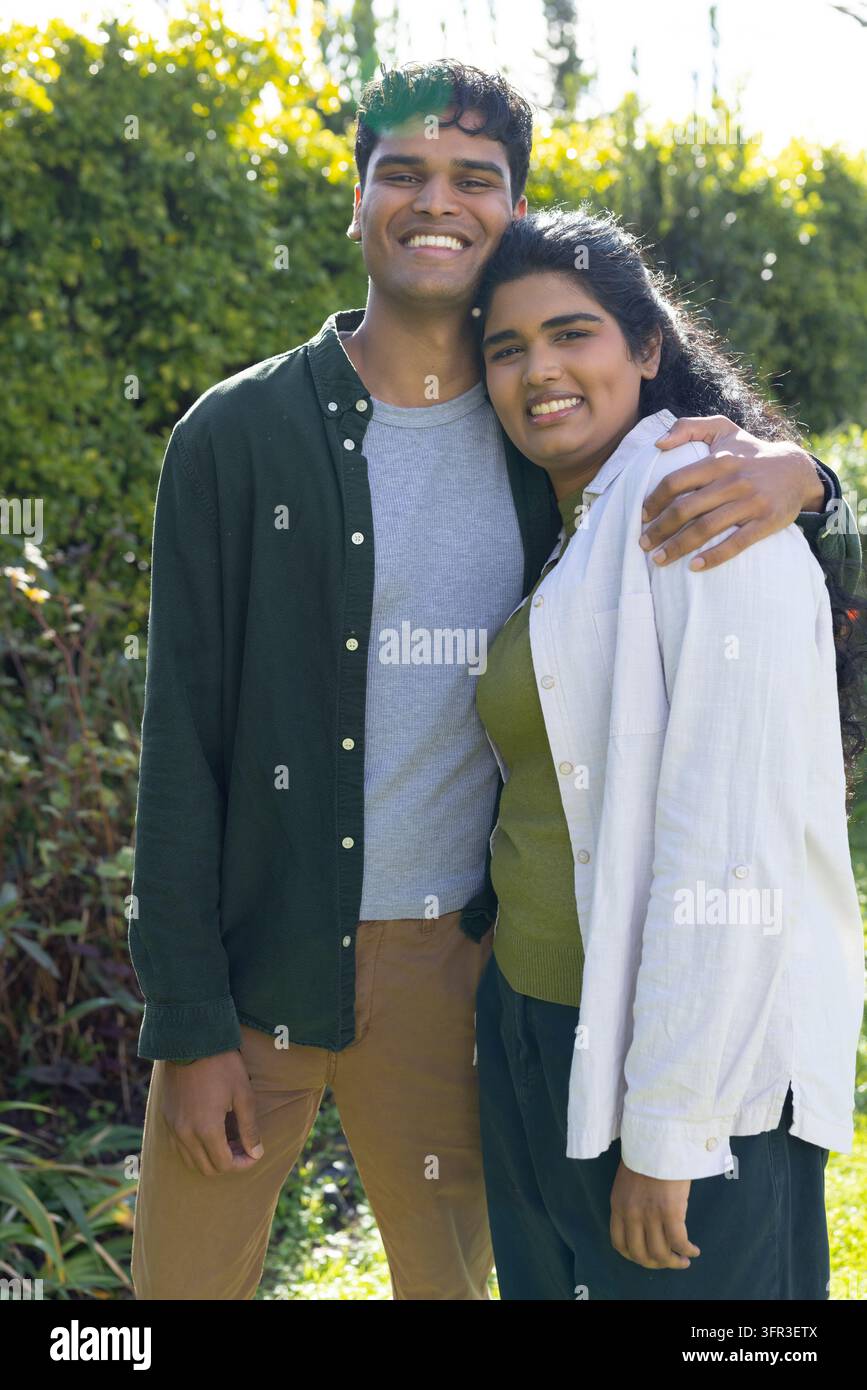 Coppia indiana in piedi in un giardino illuminato dal sole, abbracciati mentre indossano camicie oversize casual e pantaloni marrone chiaro Foto Stock