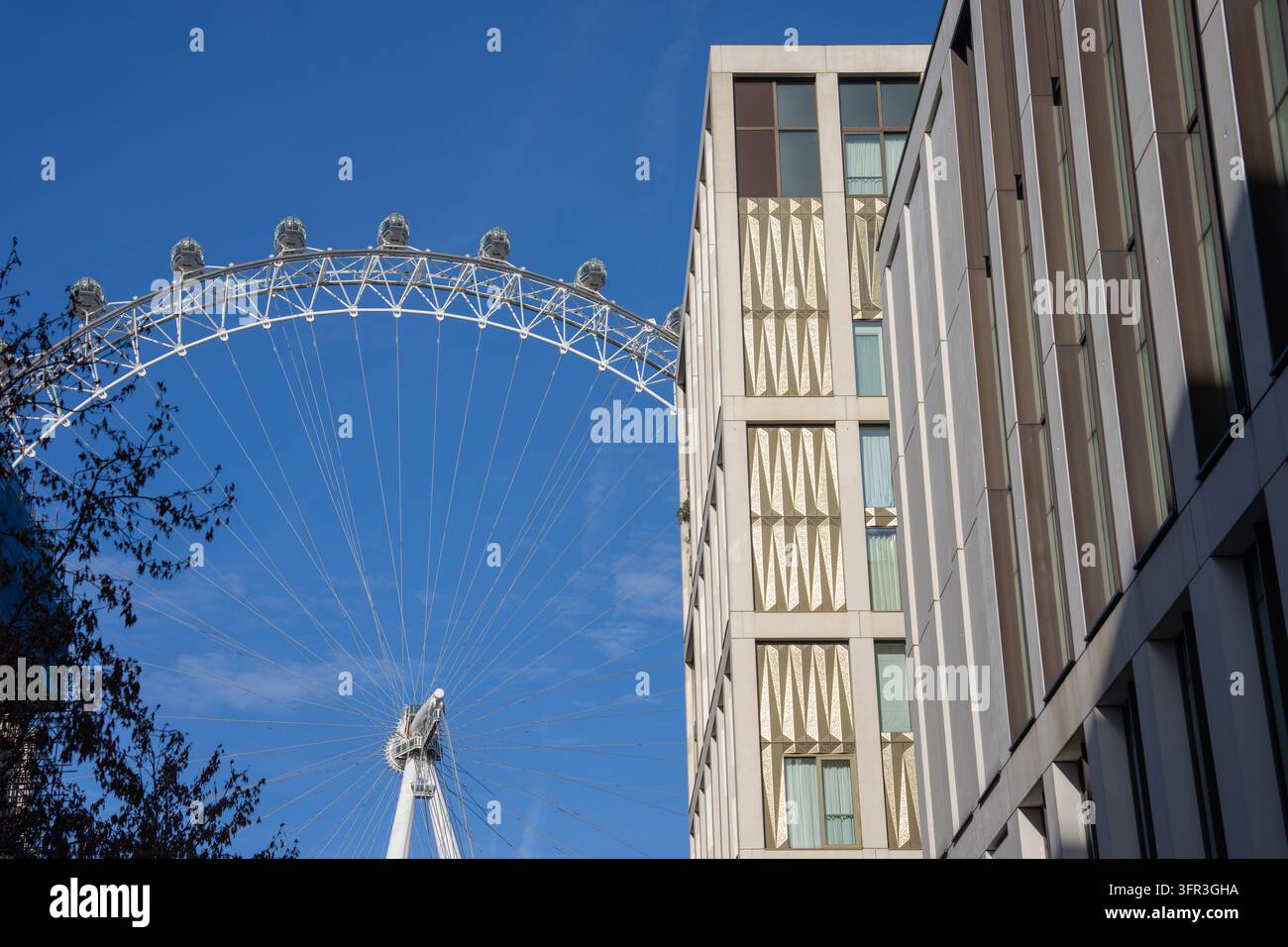 Vista della ruota panoramica iconica del London Eye incorniciata da edifici geometrici moderni di South Bank contro un cielo blu brillante. Foto Stock