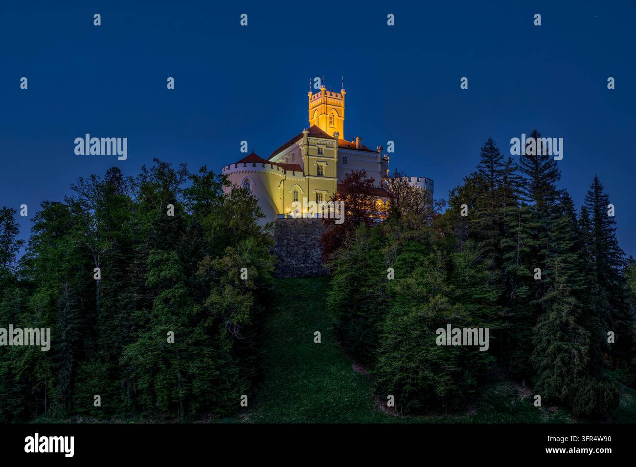 Il castello di Trakošćan illuminato, che si erge sopra una collina densamente boschiva, è illuminato al crepuscolo sotto un cielo chiaro e blu scuro. Foto Stock