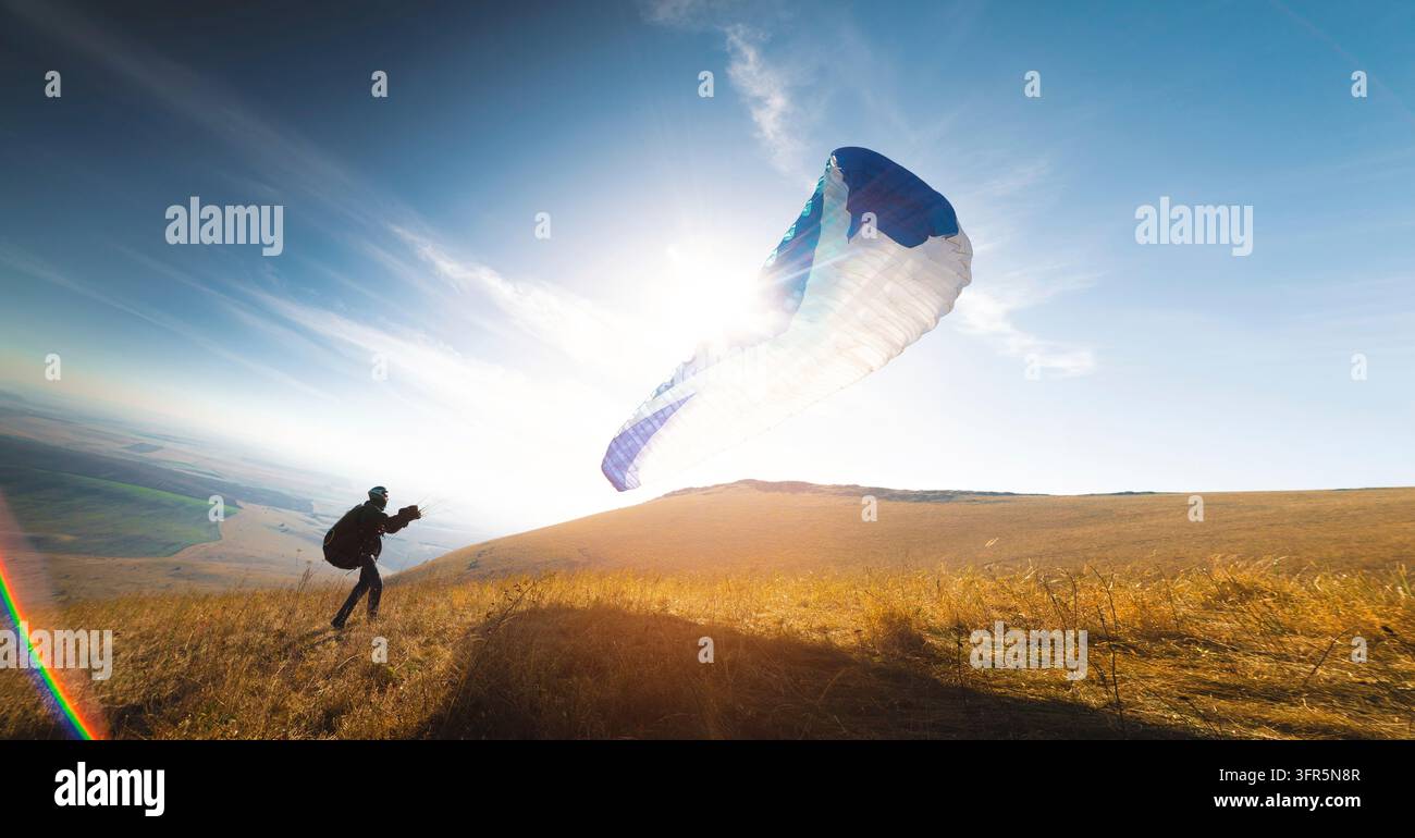Un parapendista con un paracadute blu decolla. Un uomo decolla e atterra su un campo giallo. un uomo che si prepara a decollare con un parapendio già aperto Foto Stock