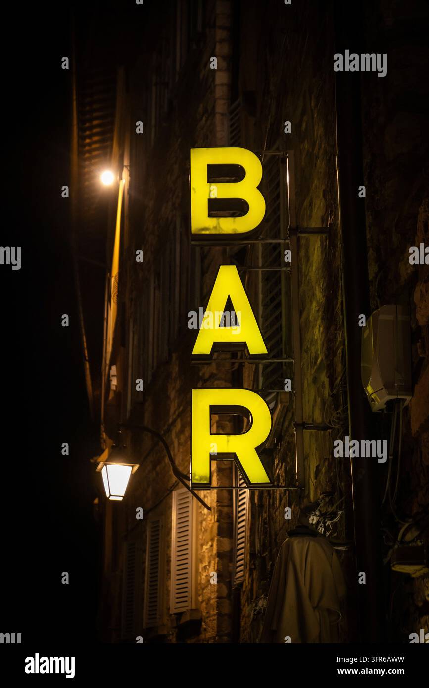Un cartello BAR giallo brilla in una stretta viuzza di notte su una vecchia facciata in pietra, Gubbio, Umbria, Italia Foto Stock