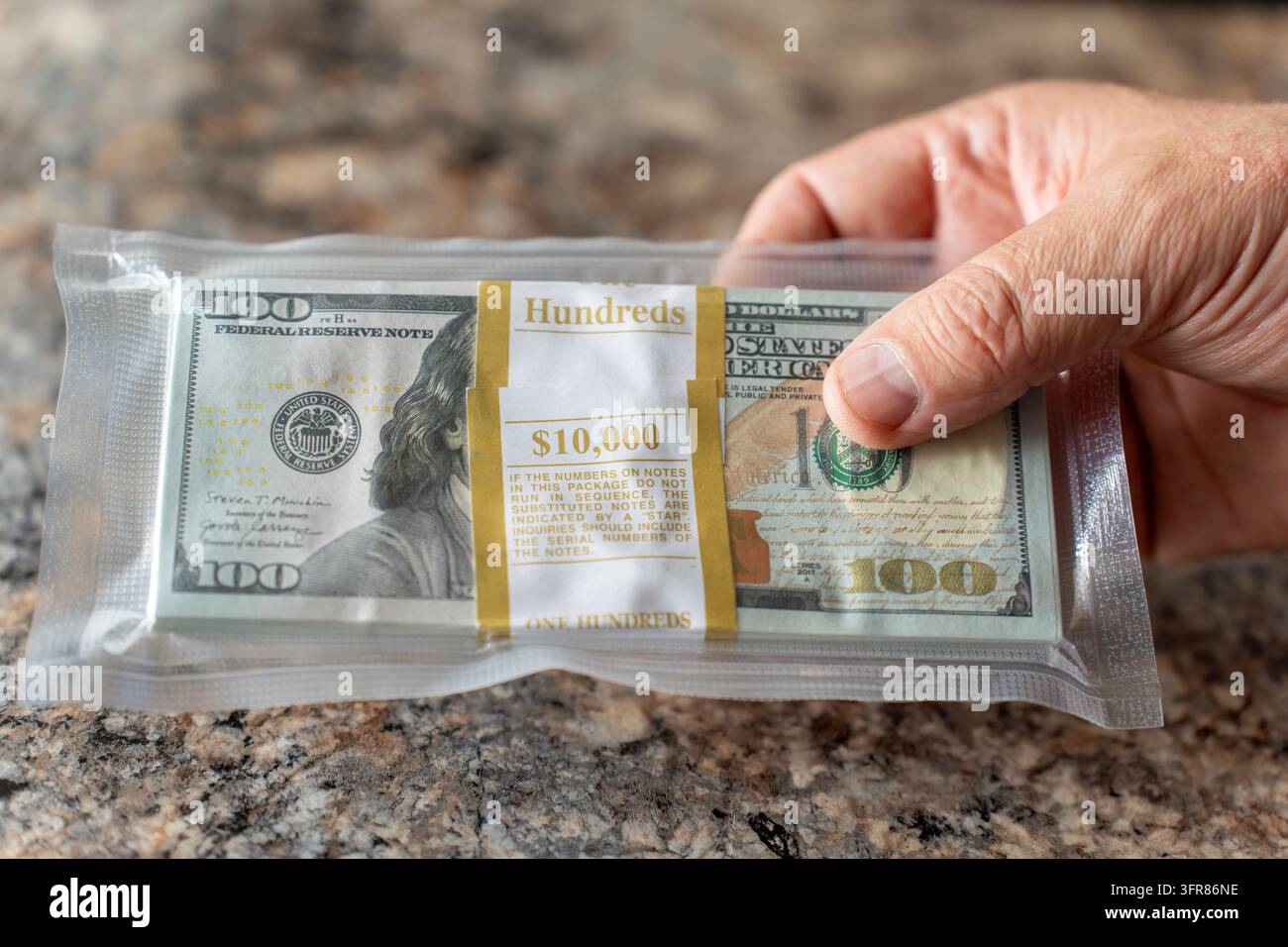 Una persona tiene una pila di banconote da cento dollari sigillate sottovuoto, per un totale di diecimila dollari, che mostra ricchezza, risparmi o una transazione importante. Conservazione impropria Foto Stock