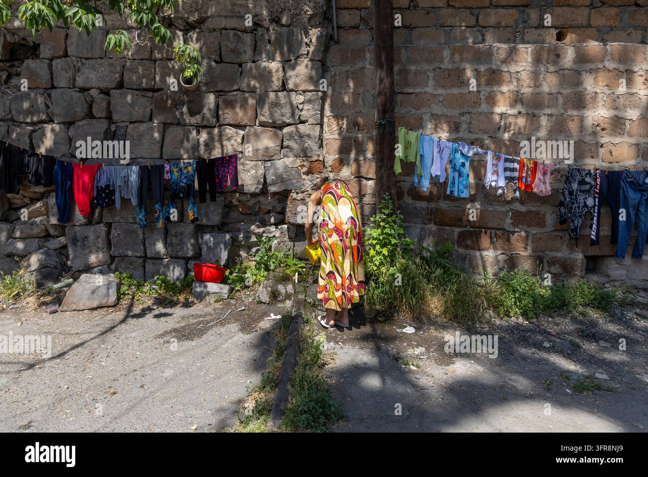 Donna china su una sorgente d'acqua per lavare i panni vicino a un muro di pietra con biancheria colorata che asciuga nelle vicinanze, Yerevan, Yerevan, Armenia Foto Stock