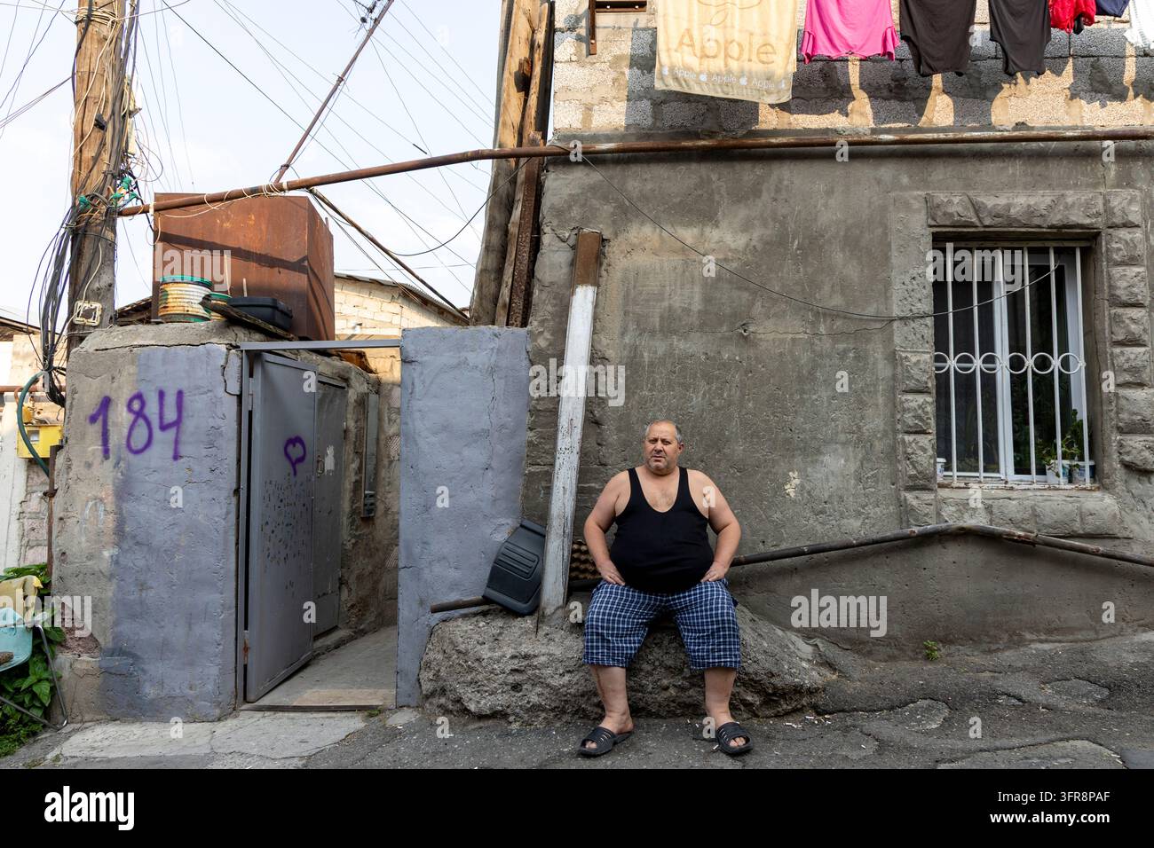 Uomo seduto fuori dalla sua casa in un modesto quartiere residenziale con il bucato appeso sopra, Yerevan, Yerevan, Armenia Foto Stock
