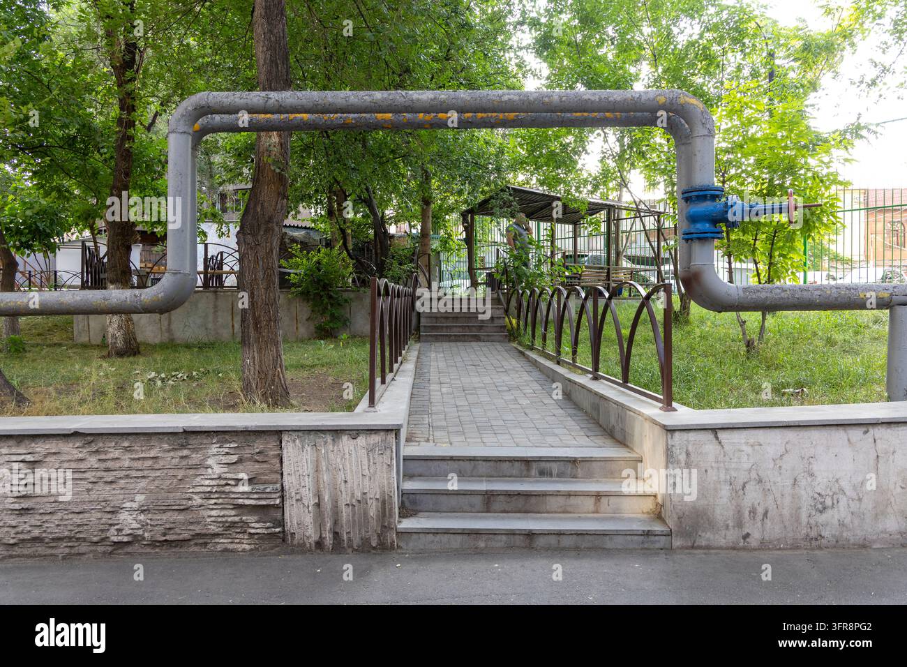 Tubazioni del sistema di riscaldamento sopraelevato che formano un arco sopra un marciapiede lastricato in un cortile verde, Yerevan, Yerevan, Armenia Foto Stock