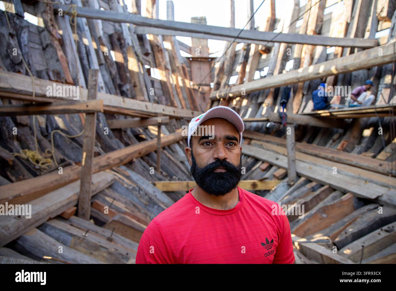 Un costruttore di navi barbuto con una camicia rossa si trova all'interno della struttura in legno di un dhow in costruzione, Gujarat, Mandvi, India Foto Stock
