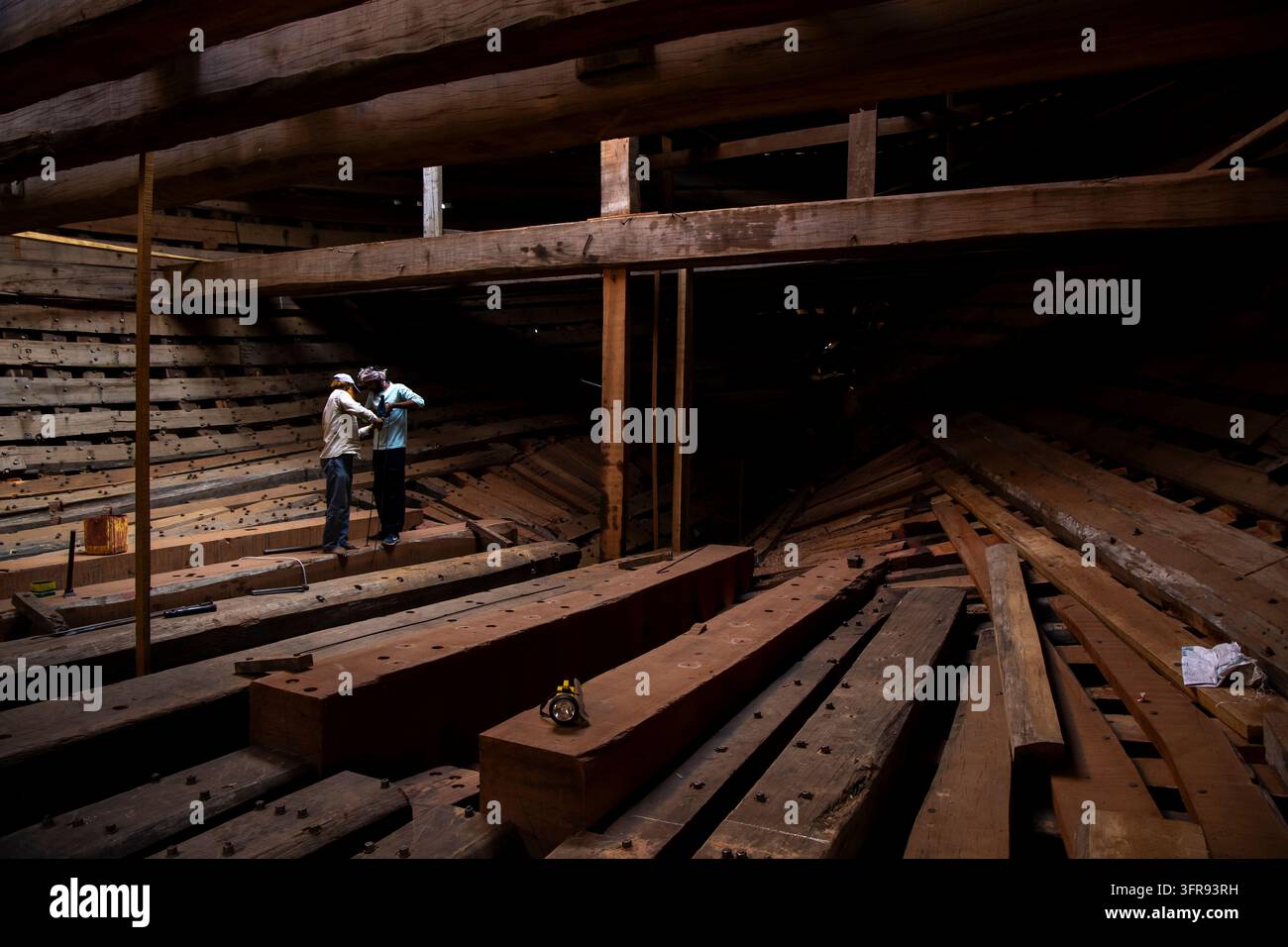 Due costruttori navali si trovano tra travi di legno all'interno dello scafo di una grande nave in costruzione in un cantiere navale a Mandvi, Gujarat, India Foto Stock