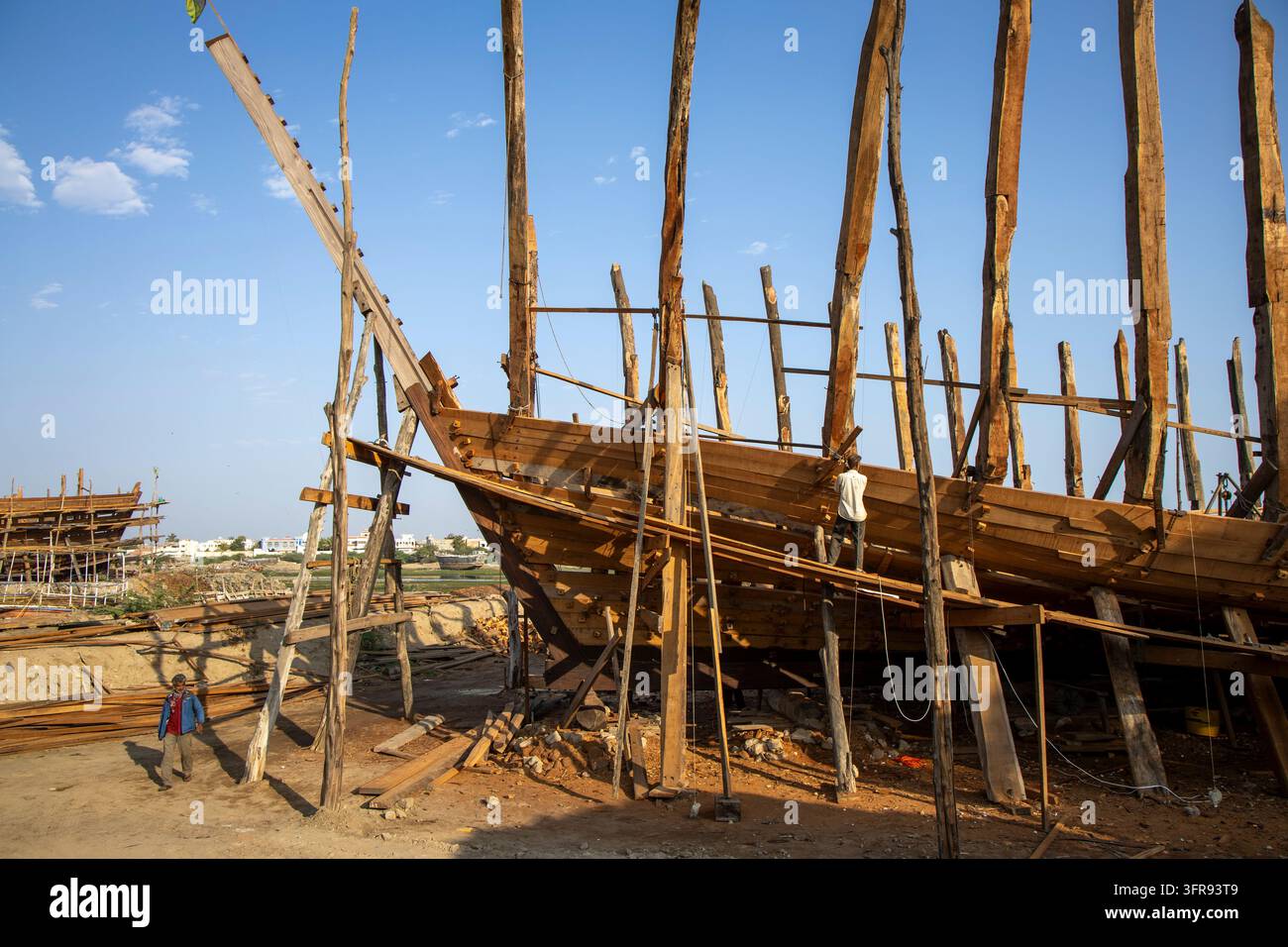 Un operaio si trova in cima allo scafo di legno di una grande nave in costruzione sostenuta da alti pali di legno, Gujarat, Mandvi, India Foto Stock