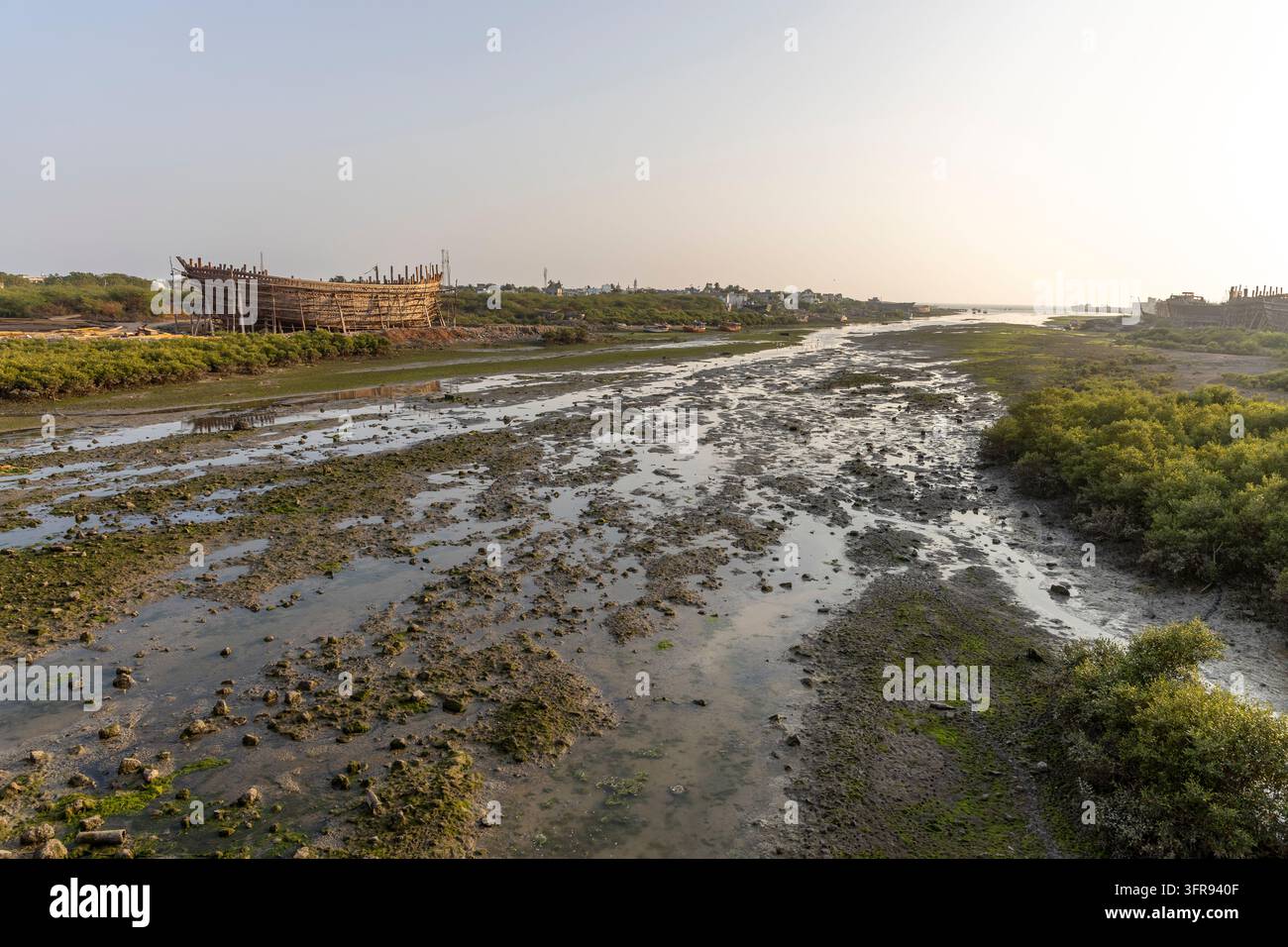 Un torrente di marea fangoso si estende verso una nave di legno in costruzione sulla riva lontana, Gujarat, Mandvi, India Foto Stock