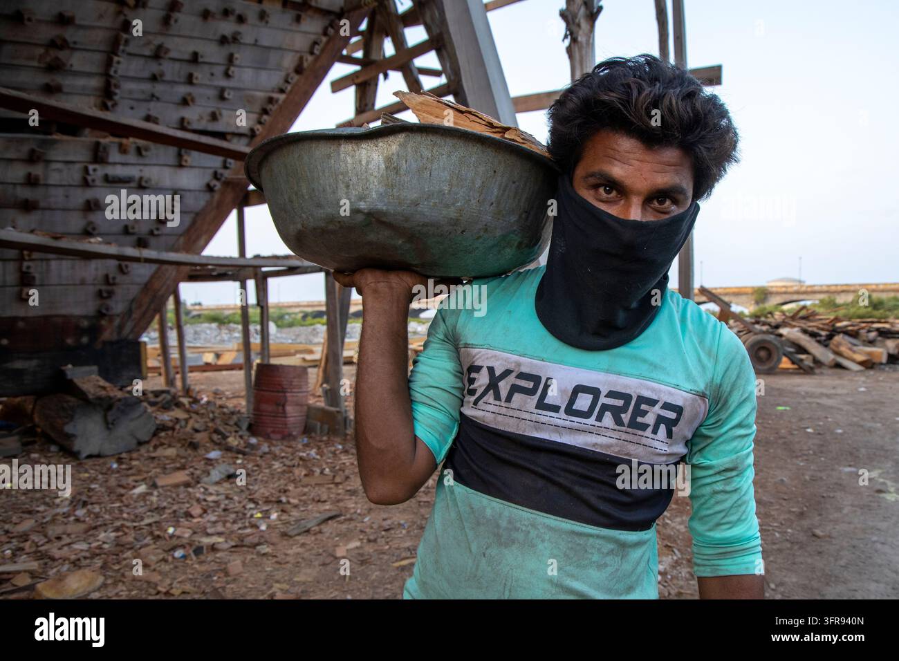 Un operaio porta una bacinella di metallo piena di scarti di legno sulla spalla vicino allo scafo di una nave in legno, Gujarat, Mandvi, India Foto Stock