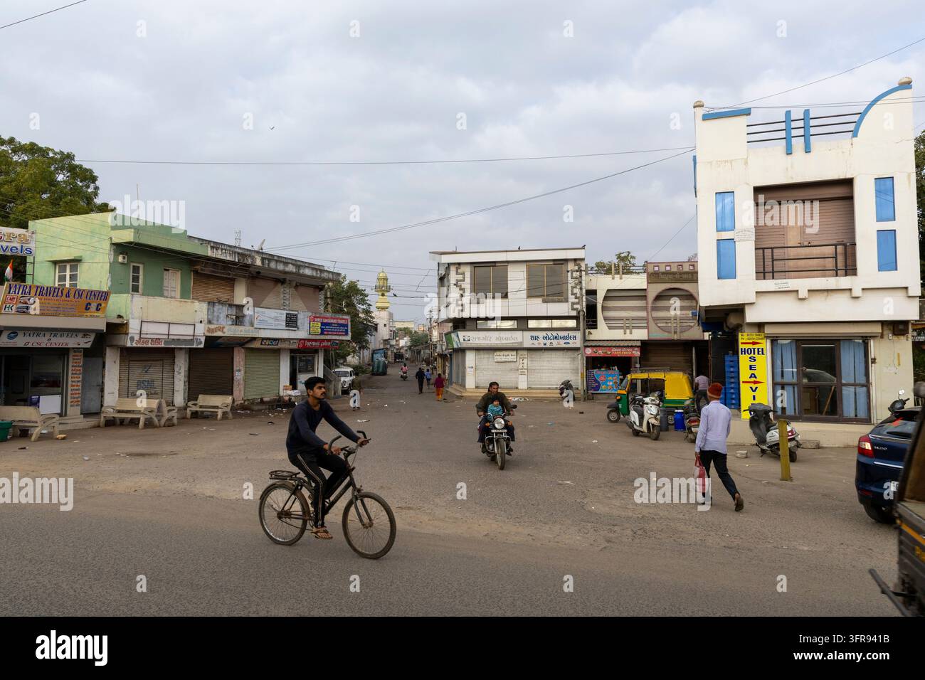 Un ciclista attraversa un incrocio tranquillo fiancheggiato da negozi chiusi in una mattina nuvolosa, Gujarat, Mandvi, India Foto Stock