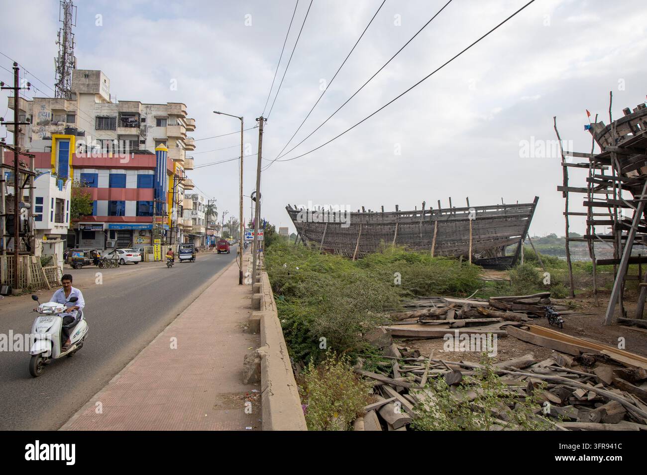 Un uomo guida uno scooter lungo una strada che si affaccia su un cantiere navale di dhow in legno dall'altra parte del fiume, Gujarat, Mandvi, India Foto Stock