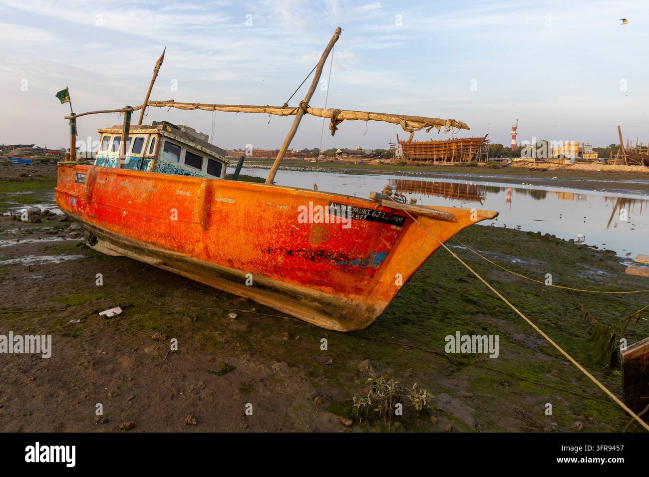 Una barca da pesca arancione è arenata su una distesa di alghe vicino a un cantiere navale e a un faro, Gujarat, Mandvi, India Foto Stock