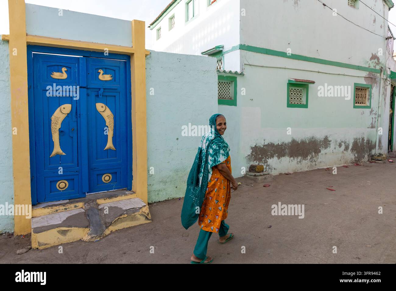 Donna sorridente con un foulard turchese che passa davanti a una porta blu decorata con motivi di pesci intagliati, Gujarat, Mandvi, India Foto Stock