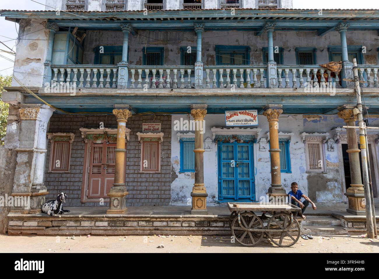 Un edificio coloniale eroso con balconi intagliati e piano terra colonnato costeggia una strada con una capra e un carretto, Gujarat, Mandvi, India Foto Stock