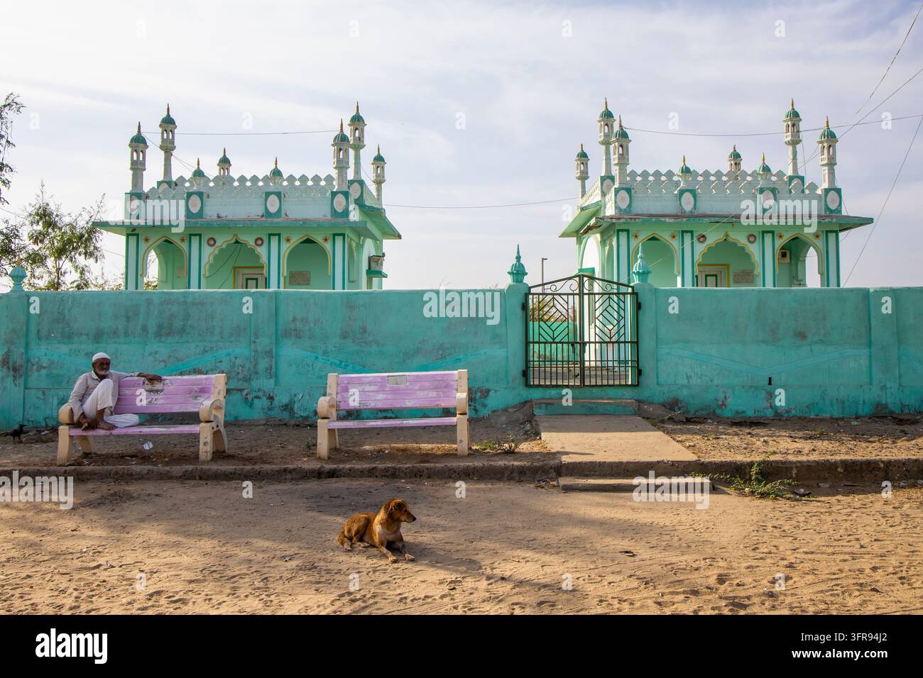 Uomo anziano che riposa su una panchina accanto a un muro turchese con due padiglioni a cupola e un cane nelle vicinanze, Gujarat, Mandvi, India Foto Stock