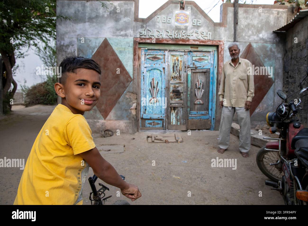 Ragazzo sorridente in bicicletta davanti a una porta blu consumata mentre un anziano osserva, Gujarat, Mandvi, India Foto Stock