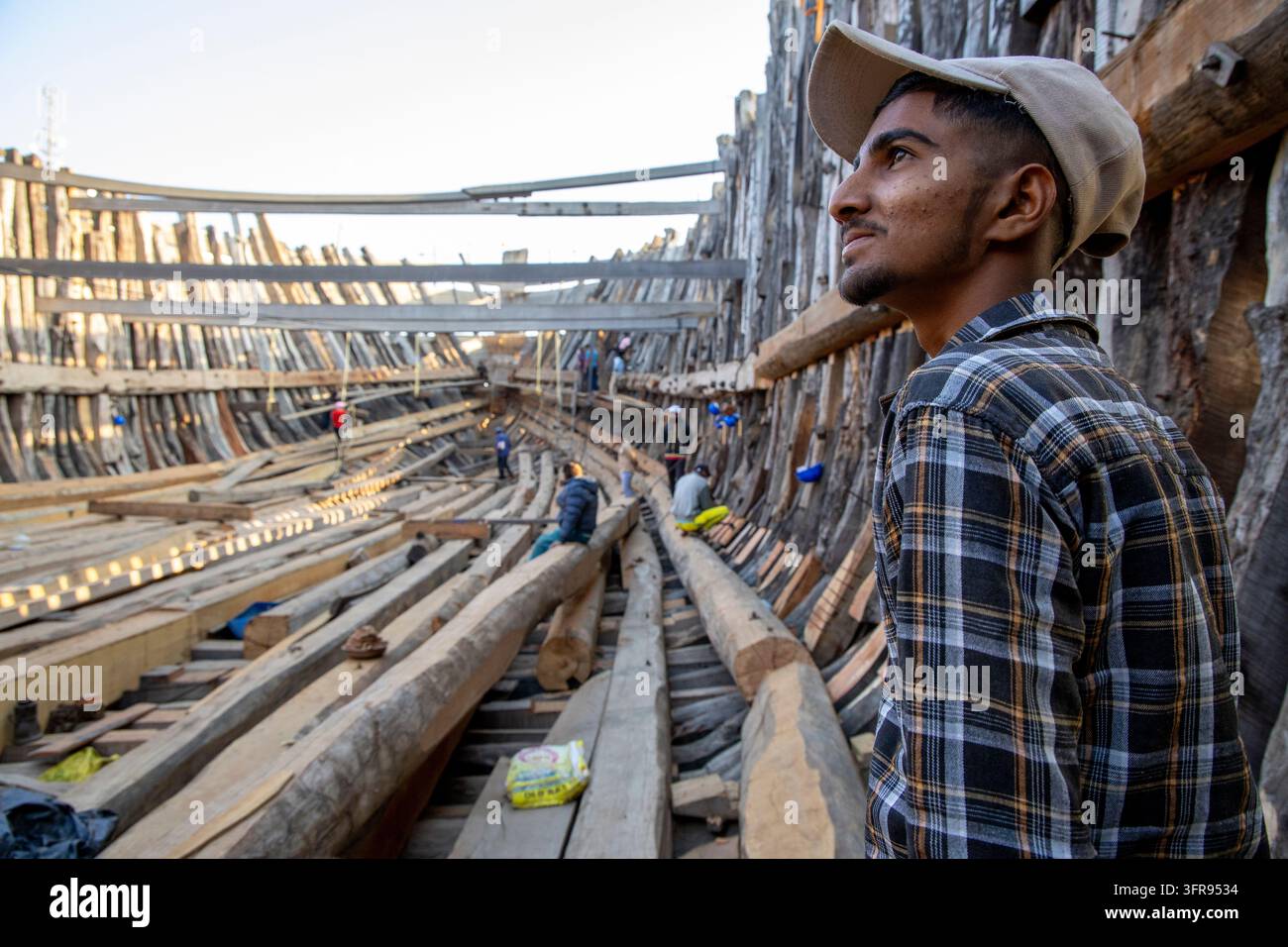 Giovane operaio di cantiere navale fissa in lontananza dall'interno dello scafo di una nave in legno in costruzione, Gujarat, Mandvi, India Foto Stock