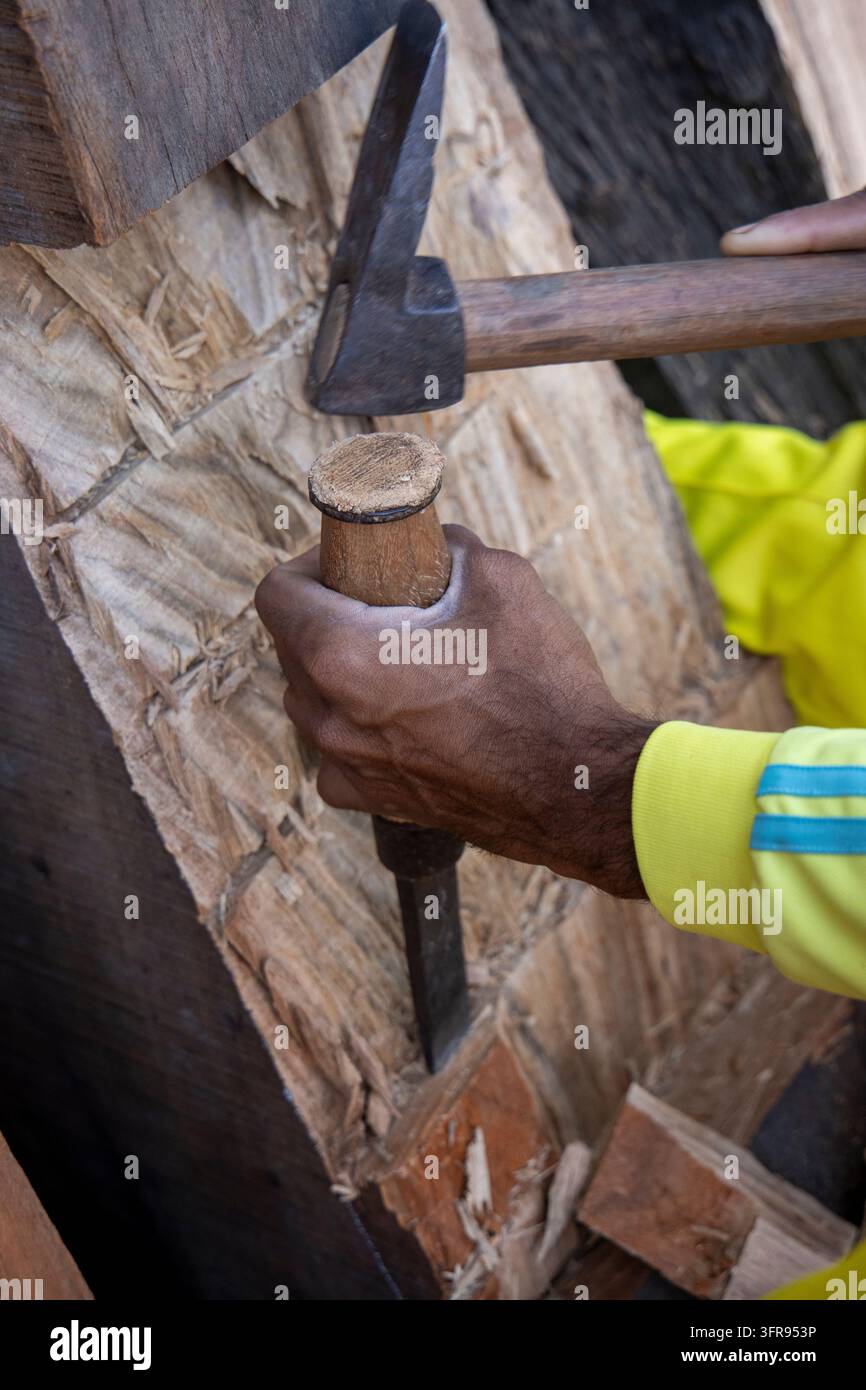 Un costruttore navale afferra con la mano un mazzuolo di legno e uno scalpello mentre modella il legname per lo scafo di una barca, Gujarat, Mandvi, India Foto Stock