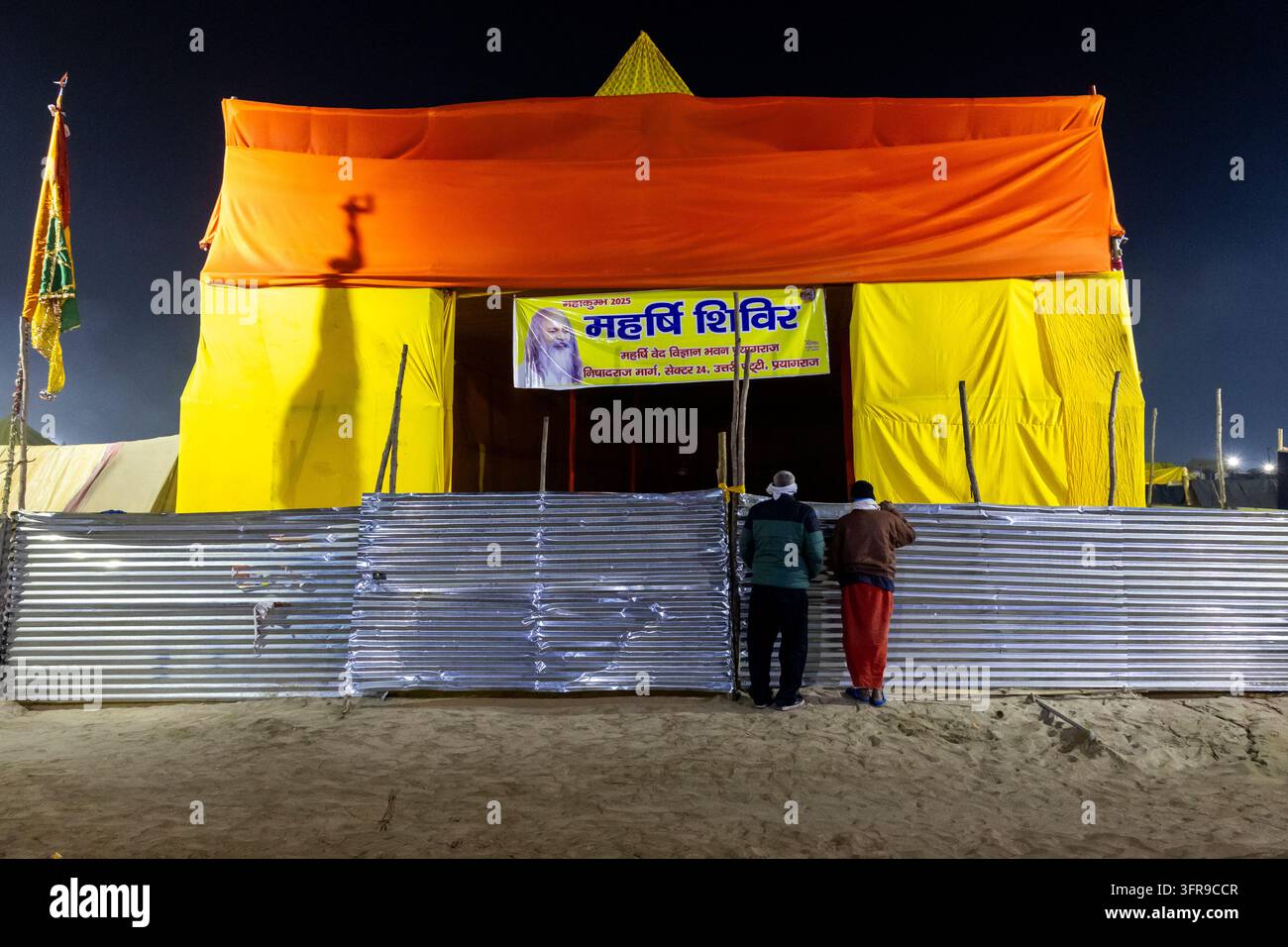 Due uomini in piedi davanti a una recinzione metallica davanti a una grande tenda arancione e gialla con uno striscione ritratto di salvia alla Maha Kumbh Mela, Uttar Pradesh, Prayagraj, India Foto Stock