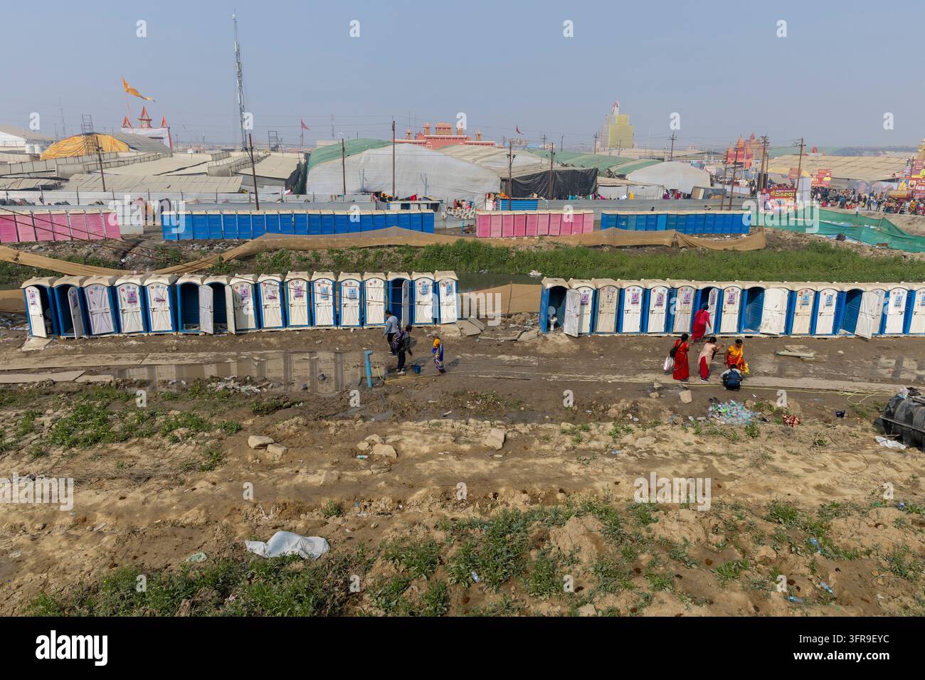 File di toilette mobili blu e bianche accanto a tende e terreno fangoso con persone che camminano in lontananza, Uttar Pradesh, Prayagraj, India Foto Stock